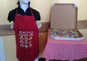 Popiersie Słowackiego w czarnej koszuli i fartuchu kuchennym a obok karton ze słodyczami.